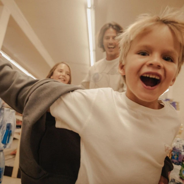 A cheerful young boy smiles broadly while being pushed in a shopping cart in a supermarket aisle, with two other people smiling behind him.