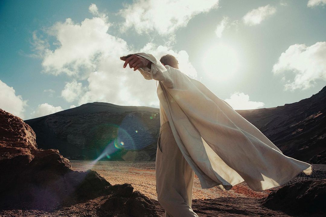 Adult in flowing white clothing stands on a rocky landscape in sunshine with cloudy skies and a mountain in the background.