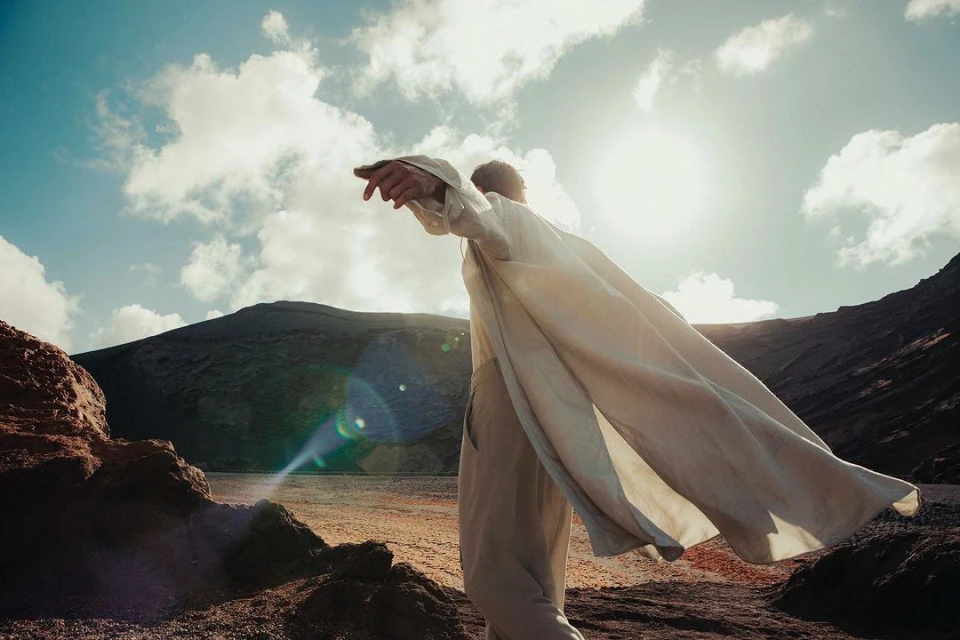 Adult in flowing white clothing stands on a rocky landscape in sunshine with cloudy skies and a mountain in the background.