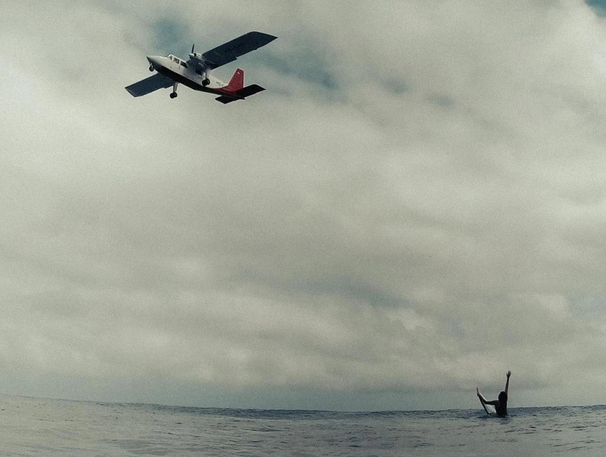 Twin-engine airplane flying above a man in the ocean with his arms raised in the air against cloudy sky, creating a striking juxtaposition.