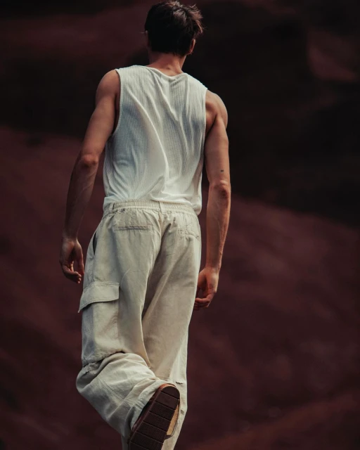 Man walking away wearing white tank top and loose beige pants against a diffused brown background. Fashion or lifestyle content.