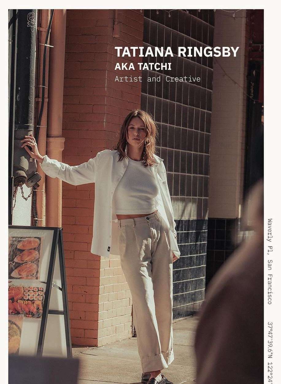 Outdoor fashion portrait of a stylish adult female in a white jacket and pants leaning against a building with an advertisement on the side.