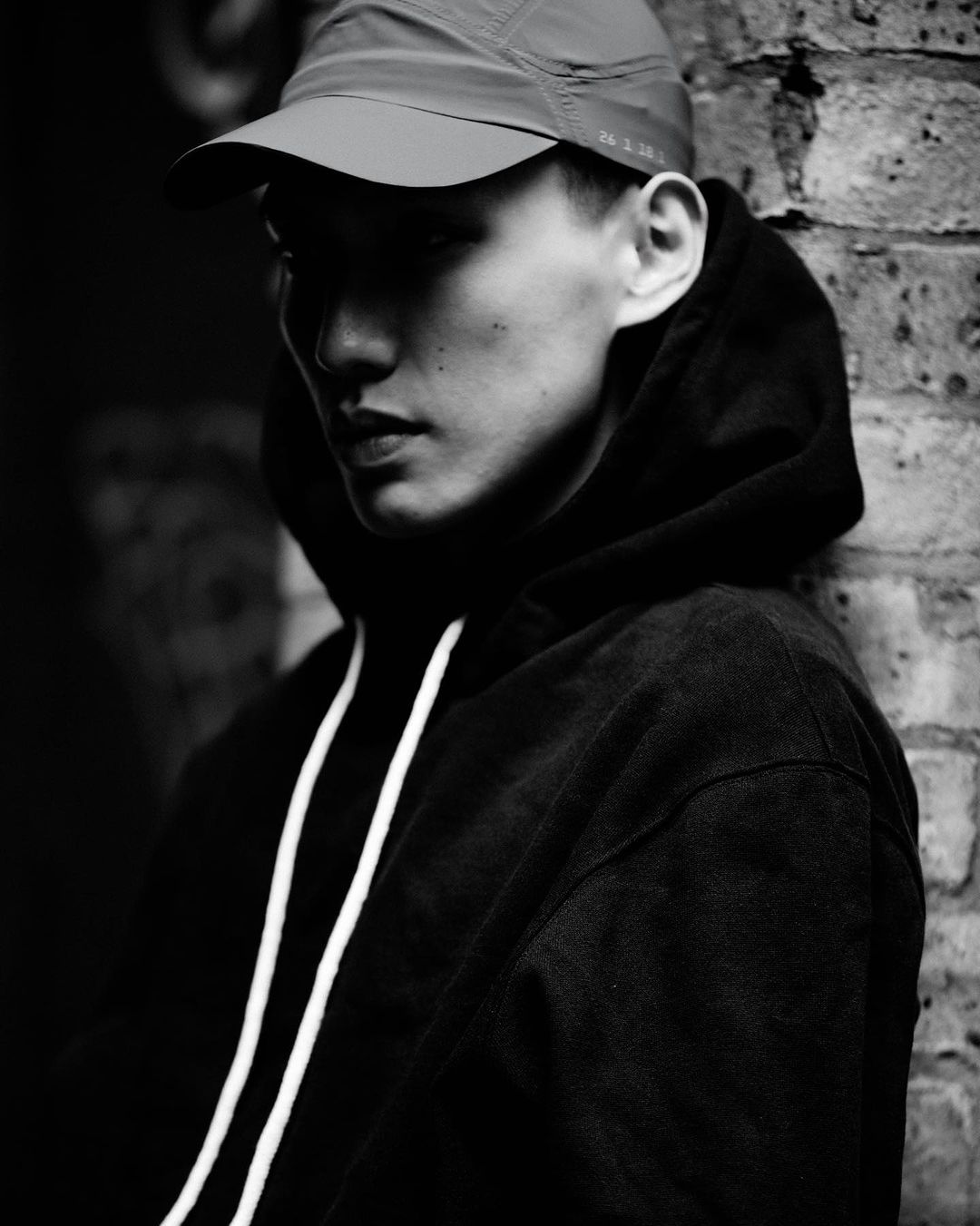 Monochrome portrait of an Asian man wearing a cap and dark hoodie leaning against a textured brick wall.