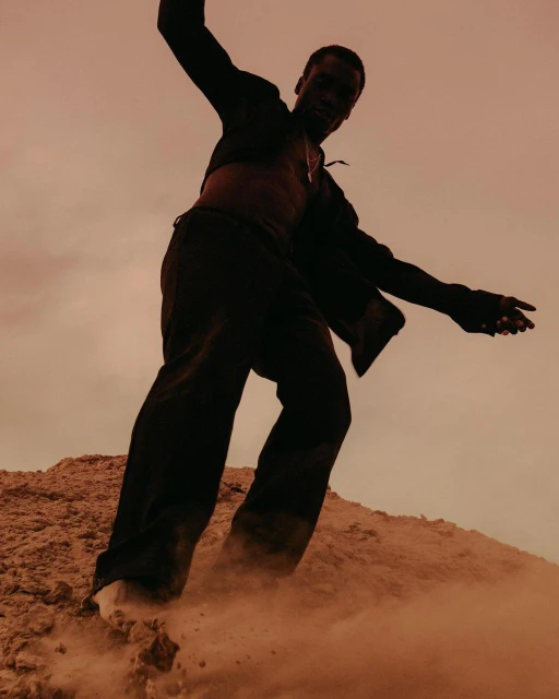 Low-angle shot of a black male model in a cropped jacket and pants posing on a sandy hill against a peach sky during daytime.