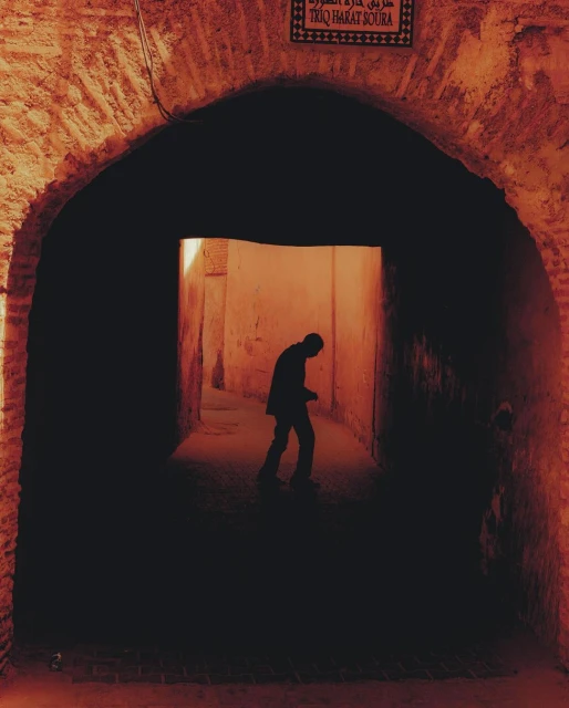 Silhouette of a man walking through an arched passageway in a historic urban area, featuring warm colors and dark shadows.