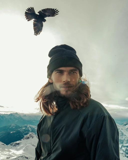 Man with long hair and a beanie stands on a snowy mountain top with a bird flying overhead, set against a light sky.