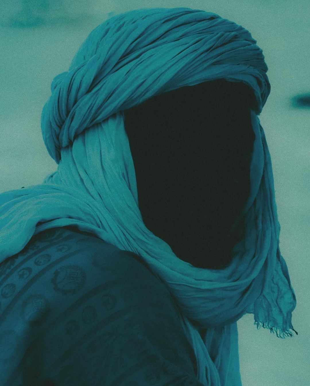 Man wearing a blue turban and patterned top stands against a desert backdrop in a teal-toned, medium shot focused on his headwear.