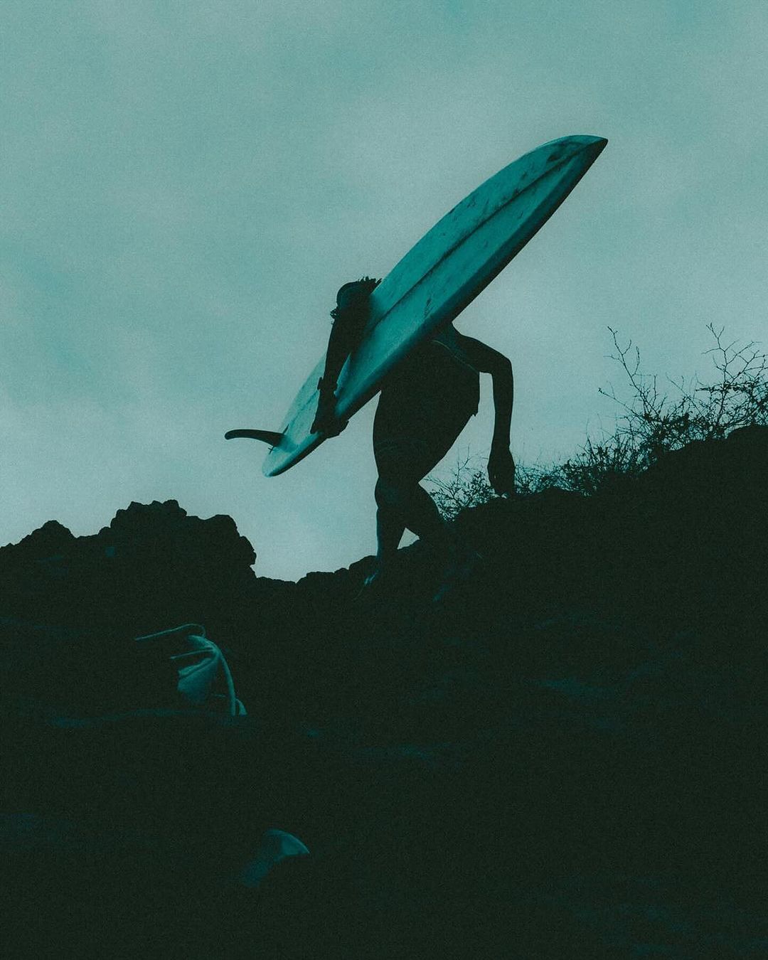 A person carrying a surfboard walks up a rocky path under a cloudy sky in a silhouette, creating a sense of adventure and athleticism.
