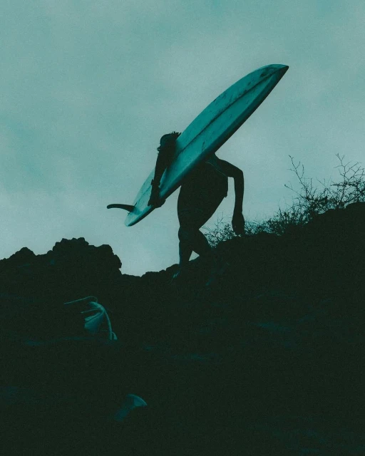 A person carrying a surfboard walks up a rocky path under a cloudy sky in a silhouette, creating a sense of adventure and athleticism.