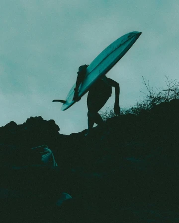A person carrying a surfboard walks up a rocky path under a cloudy sky in a silhouette, creating a sense of adventure and athleticism.