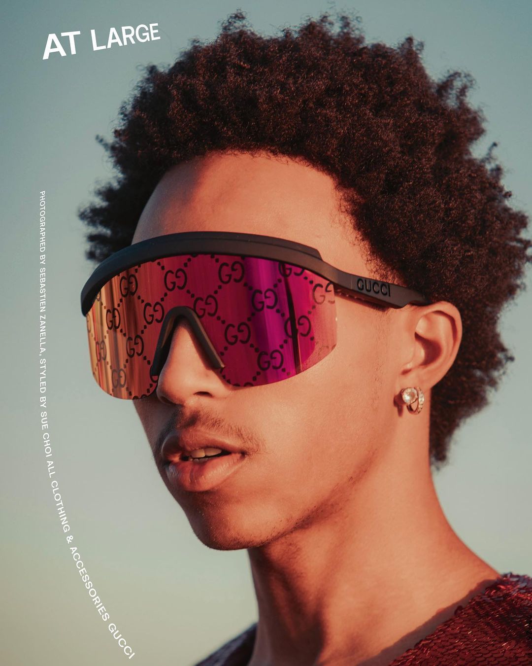 Close-up studio shot of a black male model wearing red Gucci sunglasses and an earring, set against a muted sky backdrop.