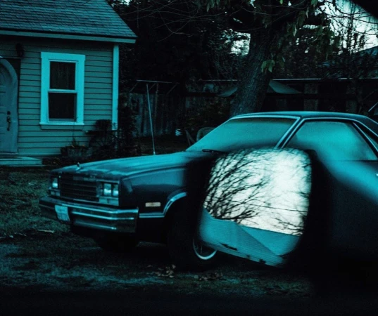 Dark teal vintage car with a reflective window parked outside a light teal siding home with trees and lawn, dusk or night scene.