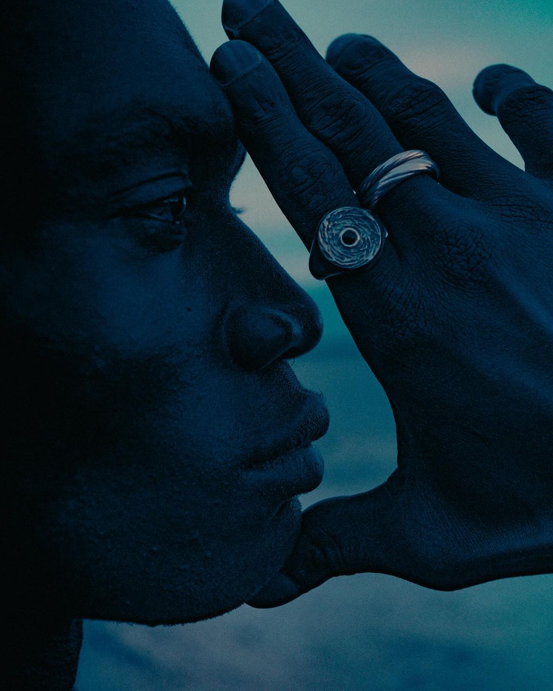 Close-up profile of a Black adult male face partially obscured by a Black hand wearing silver rings against a gradient blue sky.