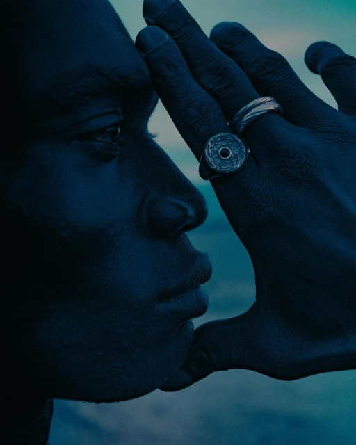 Close-up profile of a Black adult male face partially obscured by a Black hand wearing silver rings against a gradient blue sky.