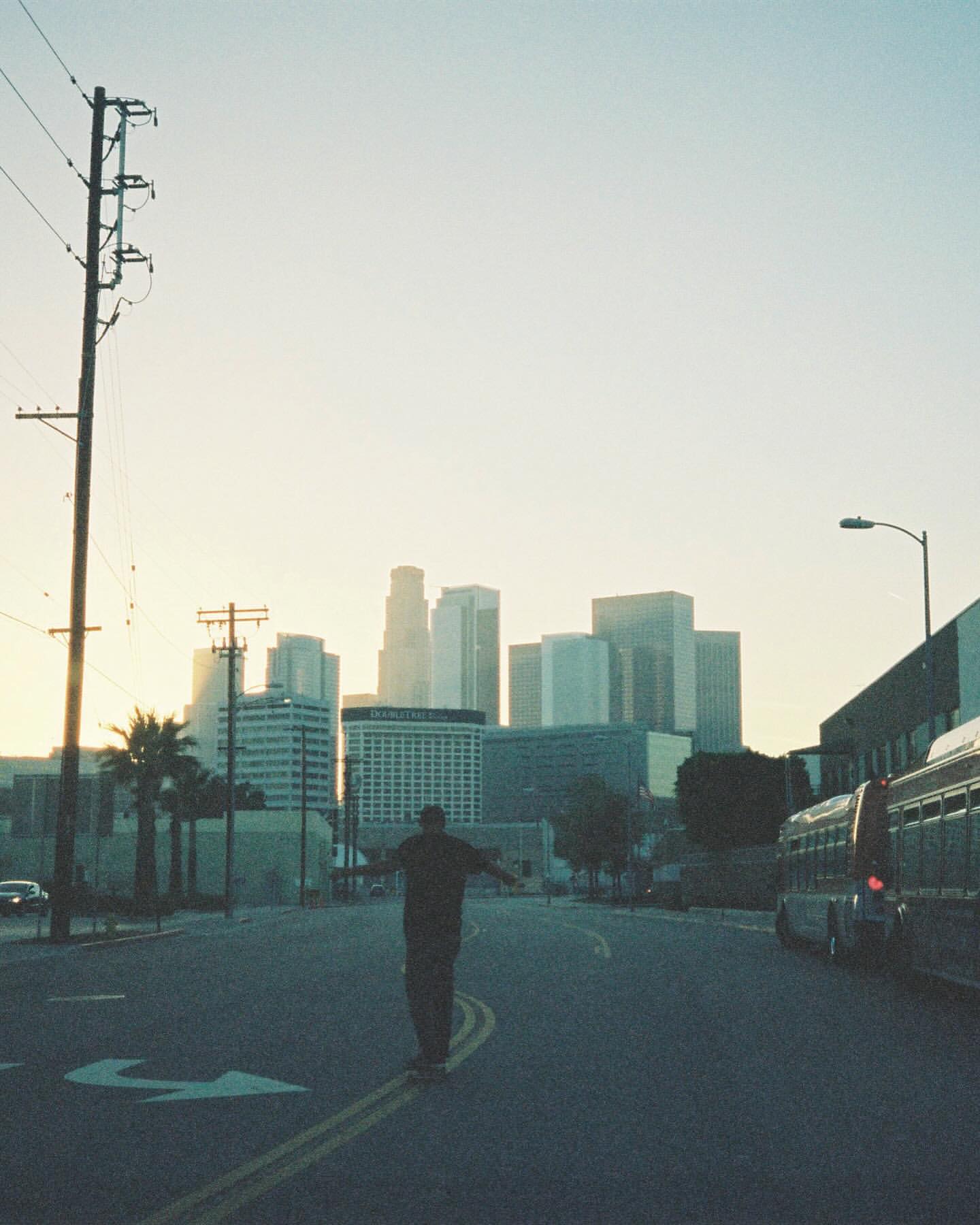 Man skateboarding on an empty street in Los Angeles with skyscrapers and city buildings in the background on a sunny day.