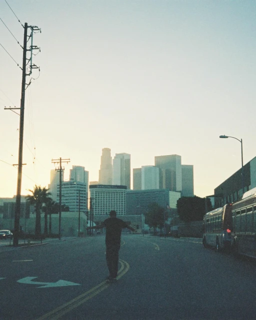 Man skateboarding on an empty street in Los Angeles with skyscrapers and city buildings in the background on a sunny day.