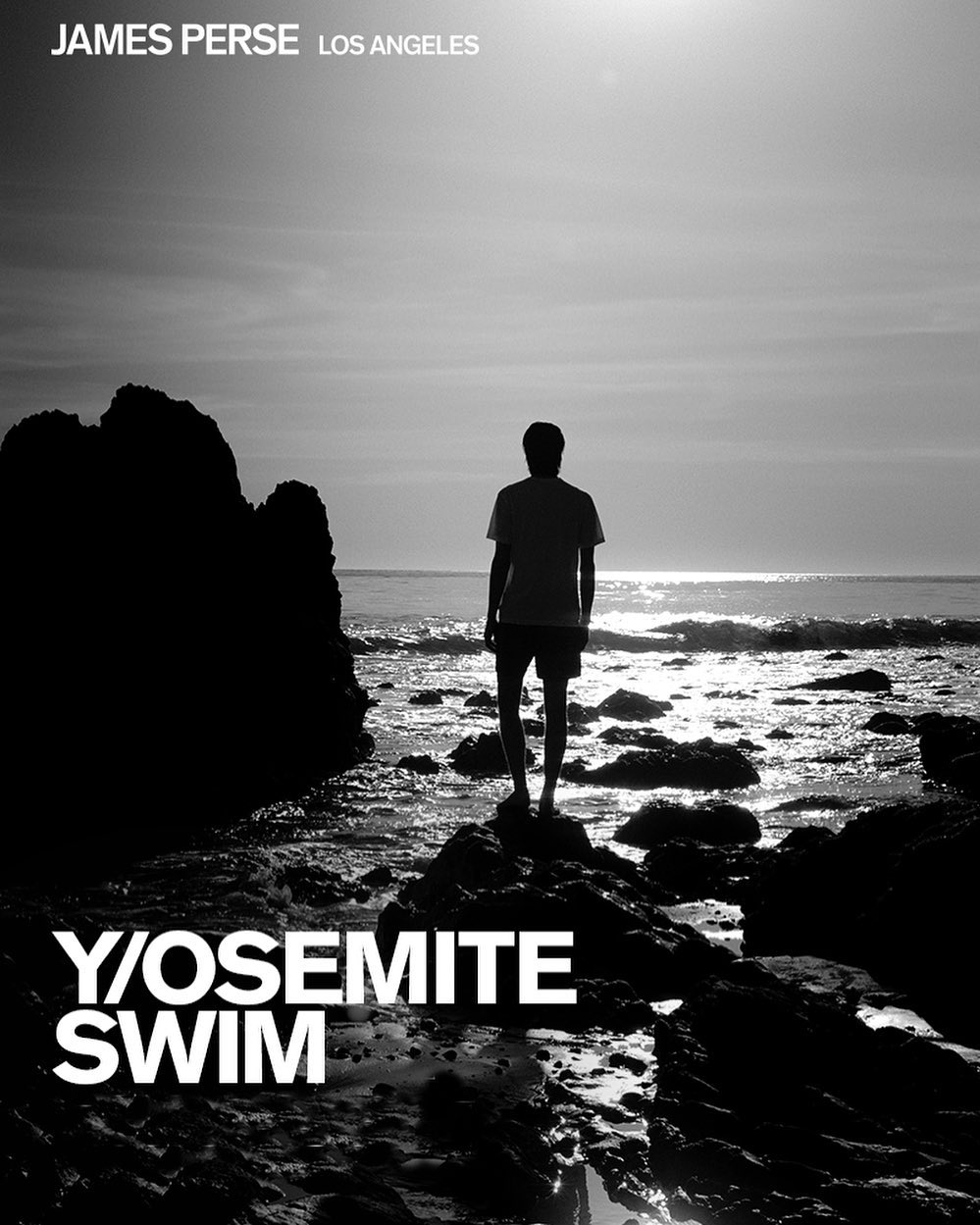 Black and white shot of a man standing on rocks at the beach, gazing out at the ocean during daytime with horizon, waves, and sky.