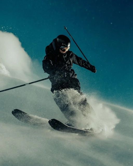 A skier in black winter gear is skiing down a snow-covered slope on a bright day, kicking up powder as they move forward, captured in motion.