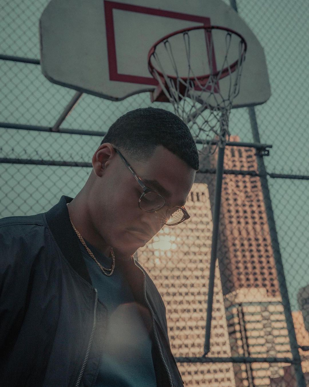 Portrait of a young man wearing glasses and jacket, standing on an urban basketball court with city buildings in the background.