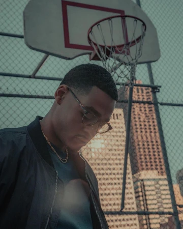 Portrait of a young man wearing glasses and jacket, standing on an urban basketball court with city buildings in the background.