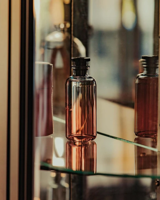 Aromatic fragrance in a glass bottle with a black cap on a glass shelf reflects in the shiny surface of a display case.