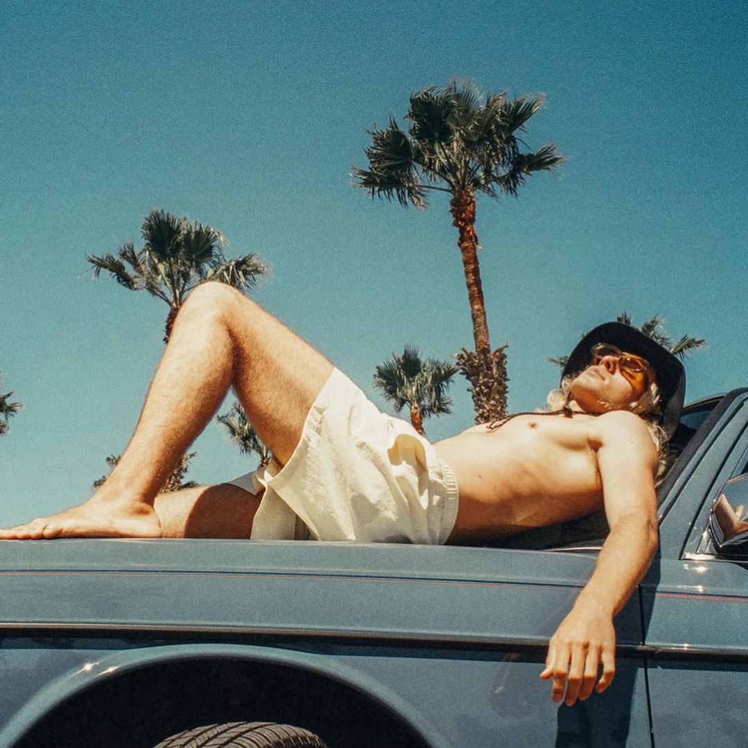 Man in shorts lies shirtless on a car hood beneath palm trees against a blue sky on a sunny day, evoking a relaxed summer vibe.