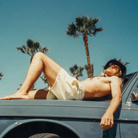 Man in shorts lies shirtless on a car hood beneath palm trees against a blue sky on a sunny day, evoking a relaxed summer vibe.