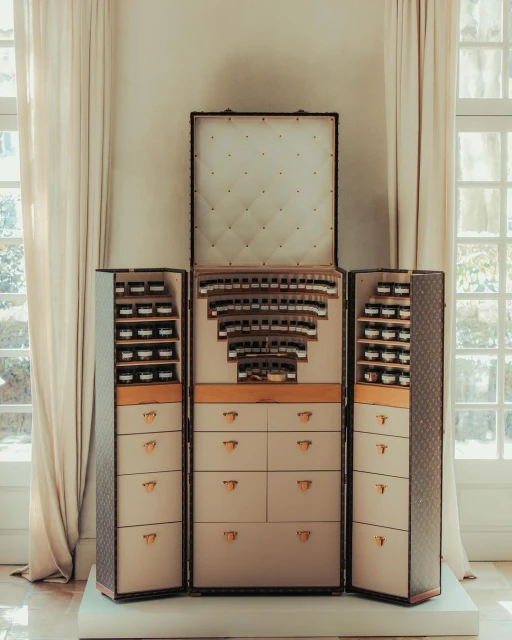 Interior of a vintage brown and beige Louis Vuitton trunk with shelving and drawers displaying a collection of perfume bottles.