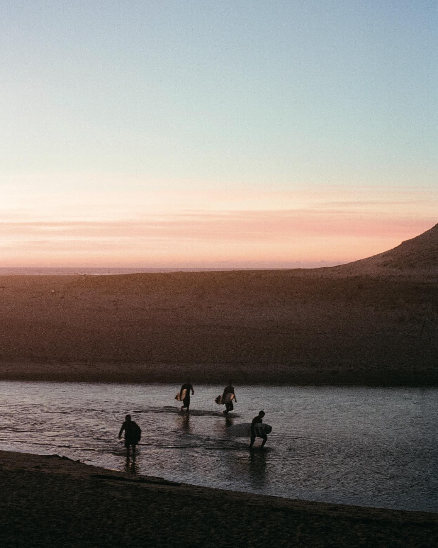 Four surfers wade through shallow water carrying surfboards at sunset, with a sandy beach and pastel sky in the background.