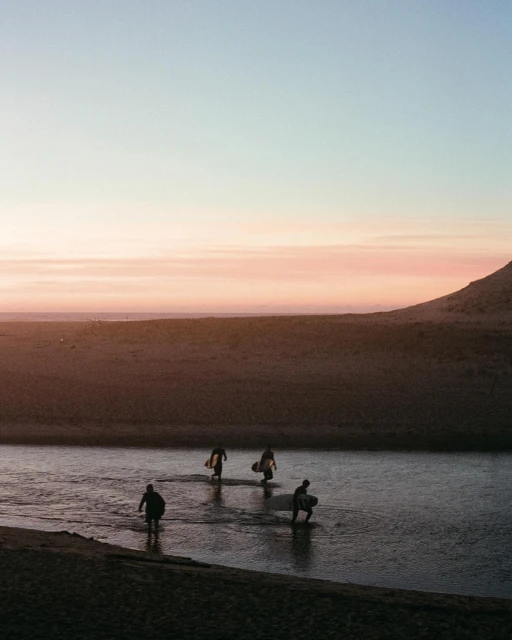 Four surfers wade through shallow water carrying surfboards at sunset, with a sandy beach and pastel sky in the background.