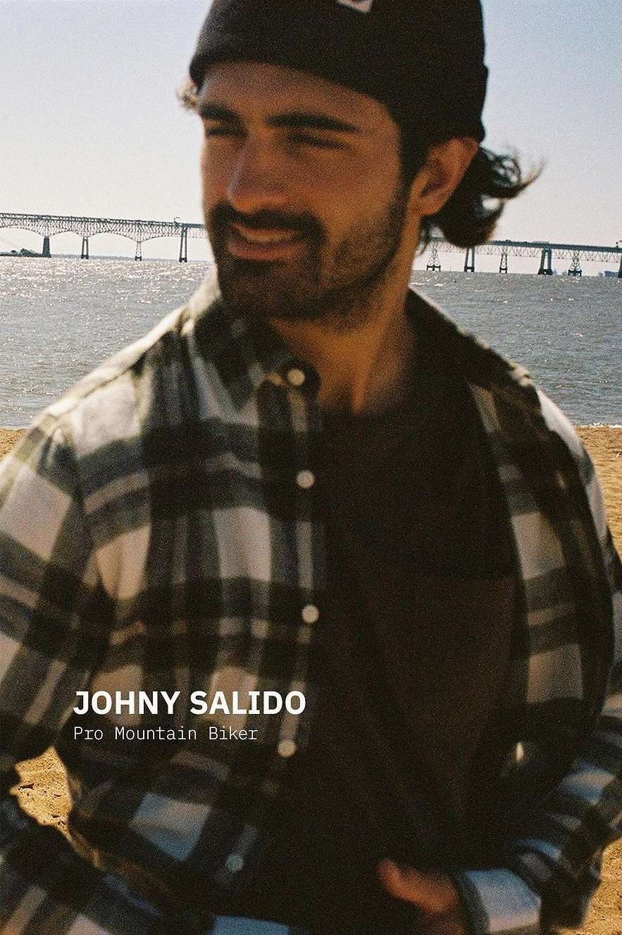 Portrait of a man in a flannel shirt and beanie, smiling with a bridge and ocean visible in the background on a sunny day.