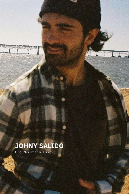 Portrait of a man in a flannel shirt and beanie, smiling with a bridge and ocean visible in the background on a sunny day.