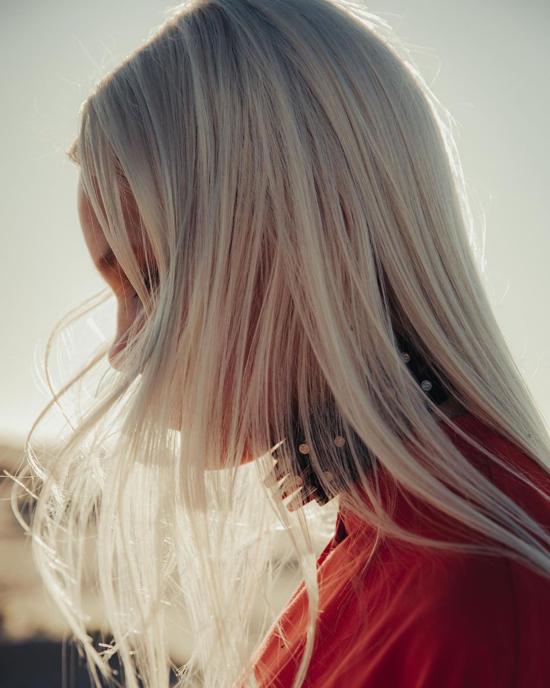 Profile view of a woman with long blonde hair wearing a red top, lit with bright and hazy sunlight, casting shadows on her neck and face.