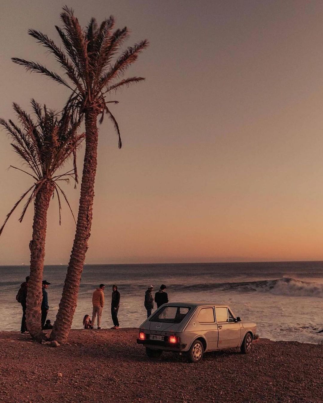 A classic car sits on a sandy beach at sunset, with palm trees and a group of people enjoying the coastal scenery.