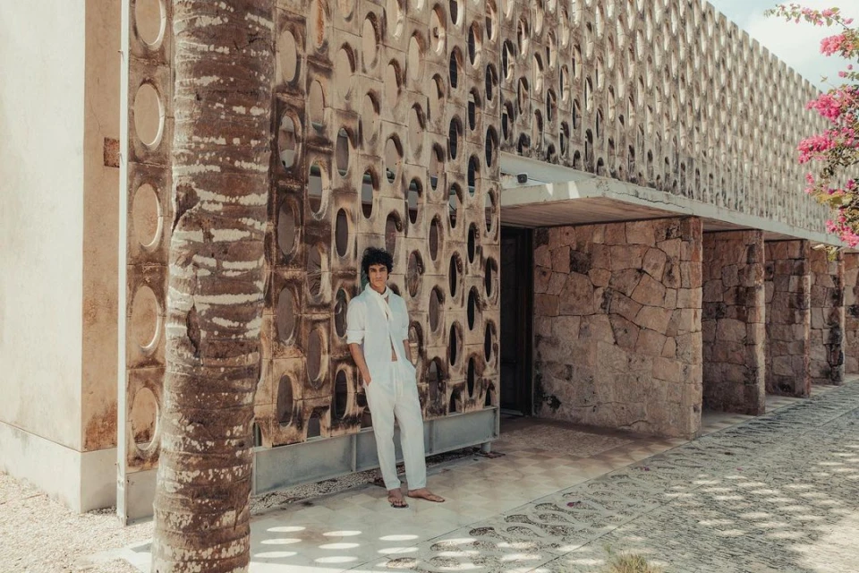 Adult male in white clothing poses in front of modern architectural building with patterned facade and palm trees outside on a sunny day.