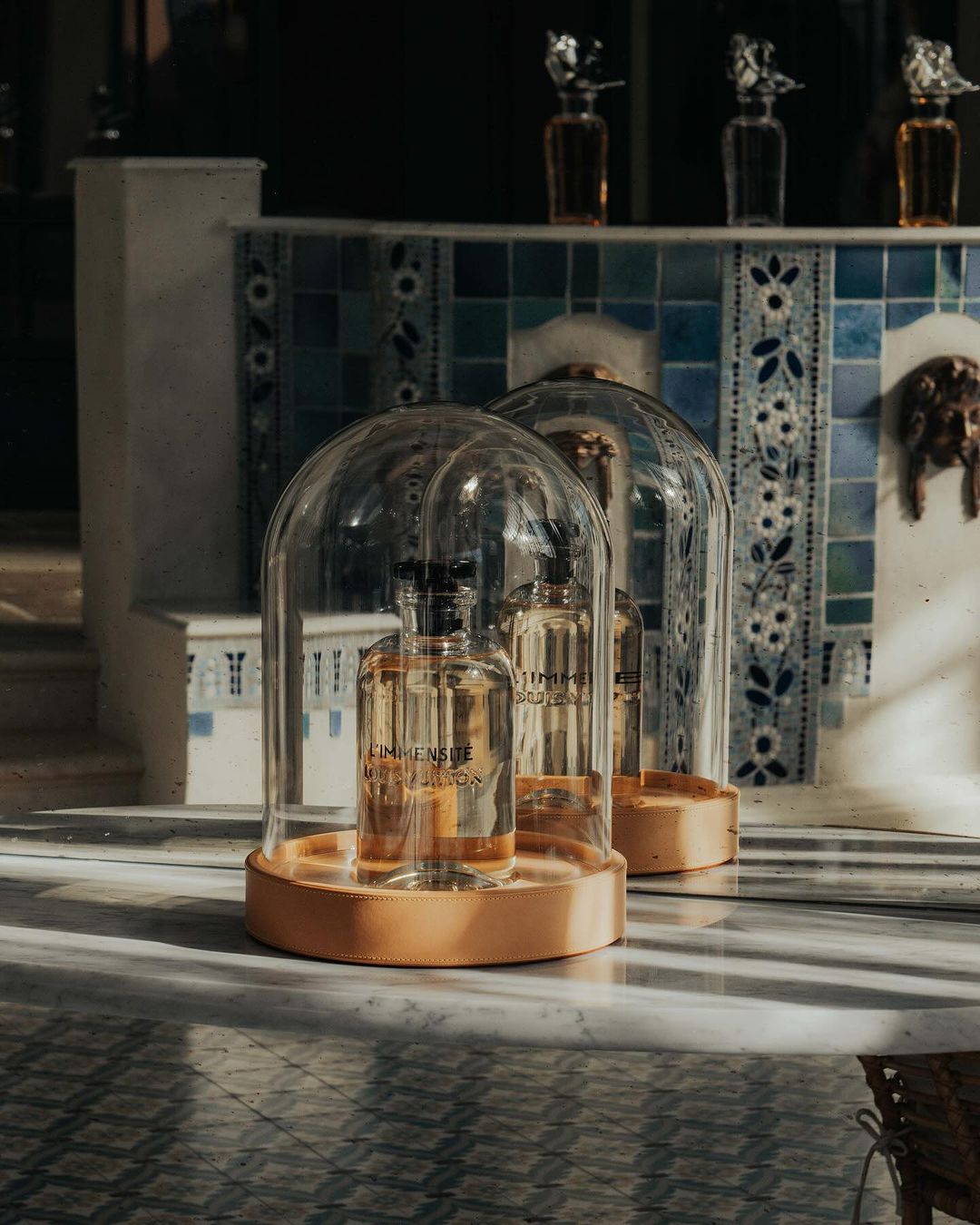 Two glass cloches containing fragrance bottles sit on a marble surface, with a tiled wall and other bottles in the soft-lit background.