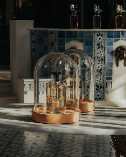 Two glass cloches containing fragrance bottles sit on a marble surface, with a tiled wall and other bottles in the soft-lit background.