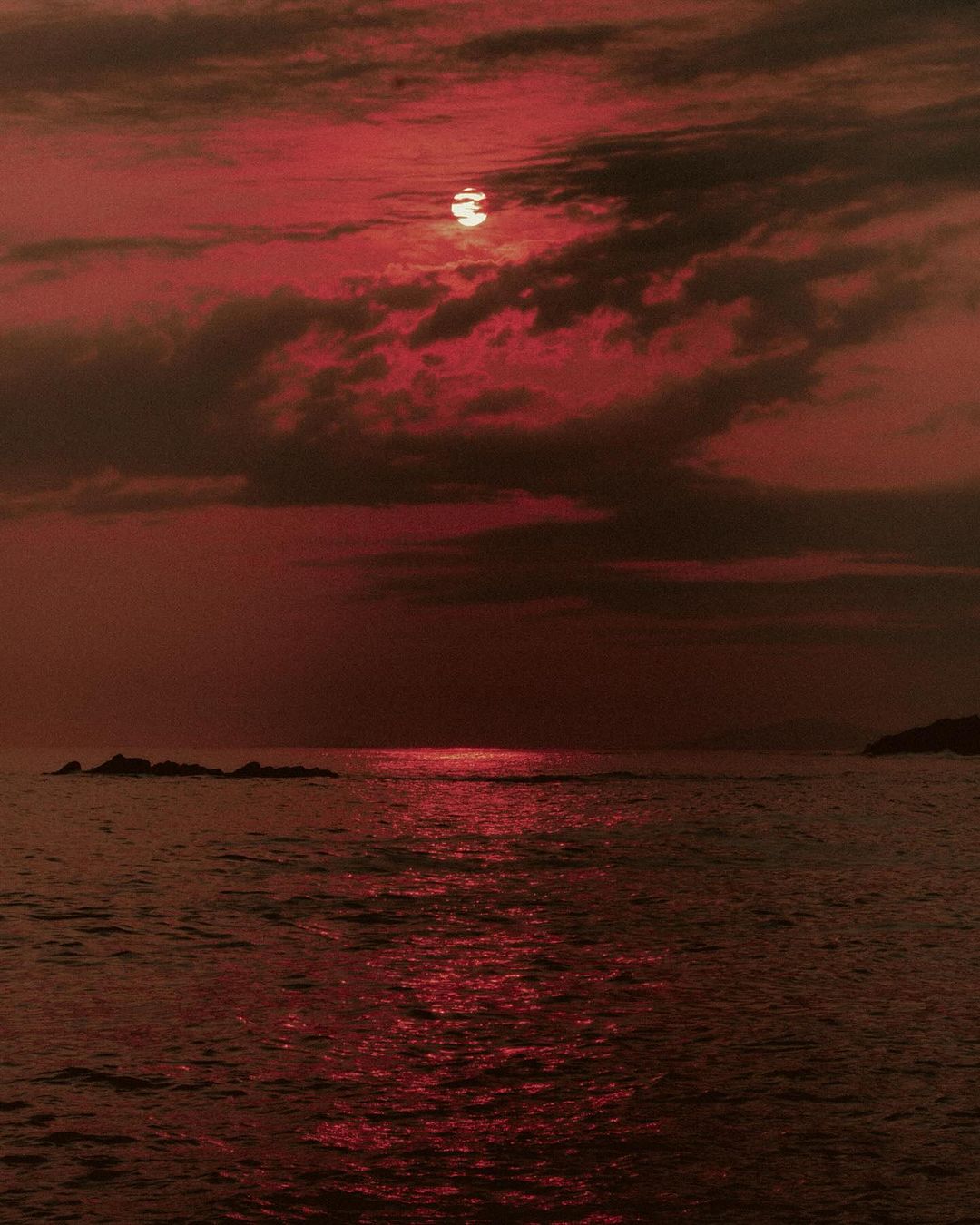 Monochromatic red scenic of the sun setting over the ocean with dark storm clouds and calm water reflecting the light in the evening.