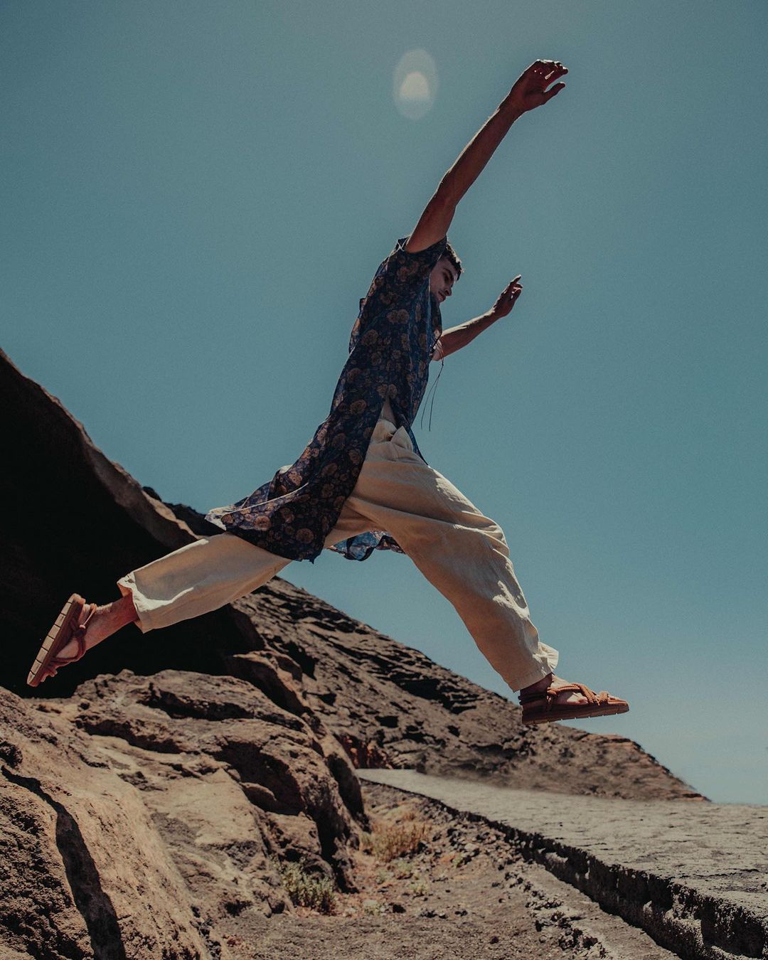 A person in ethnic attire jumps from a rocky plateau against a clear sky, capturing a dynamic moment in an arid, outdoor setting.
