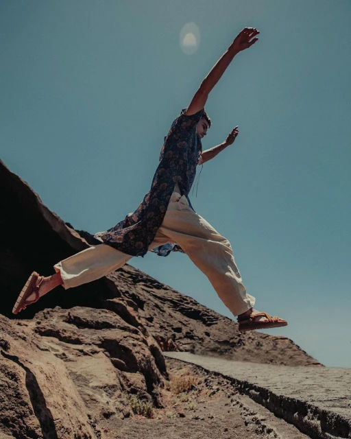 A person in ethnic attire jumps from a rocky plateau against a clear sky, capturing a dynamic moment in an arid, outdoor setting.