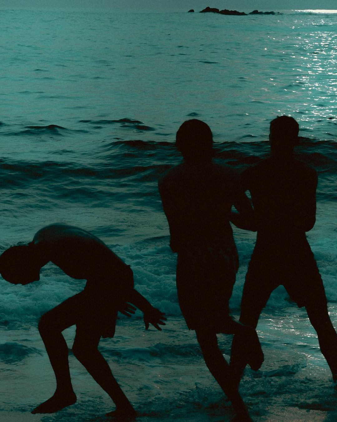 Three silhouetted figures stand on a beach under a dark sky, with the ocean stretching into the distance, creating a monochrome, moody scene.