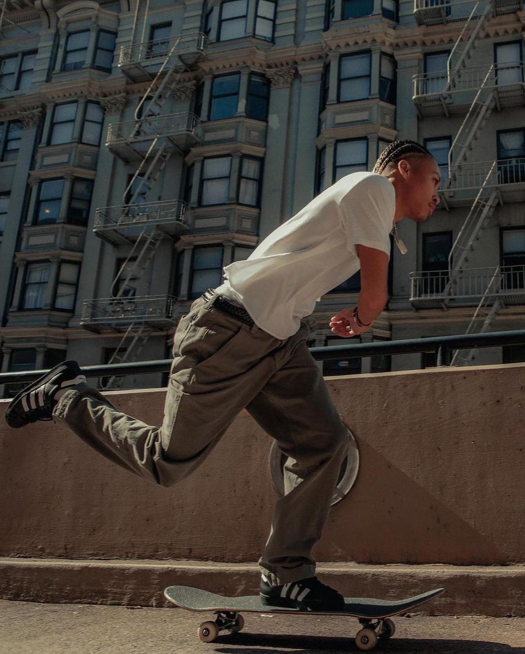 Young man skateboarding in urban environment on sunny day with city buildings in the background, wearing casual clothing and sneakers.