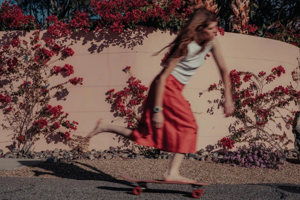 Woman in skirt skateboarding barefoot on pavement next to pink wall with red flowers in sunny outdoor setting during daytime.
