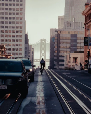 Man rides bicycle along city street with tall buildings and the Bay Bridge in the background on a sunny day in an urban environment.
