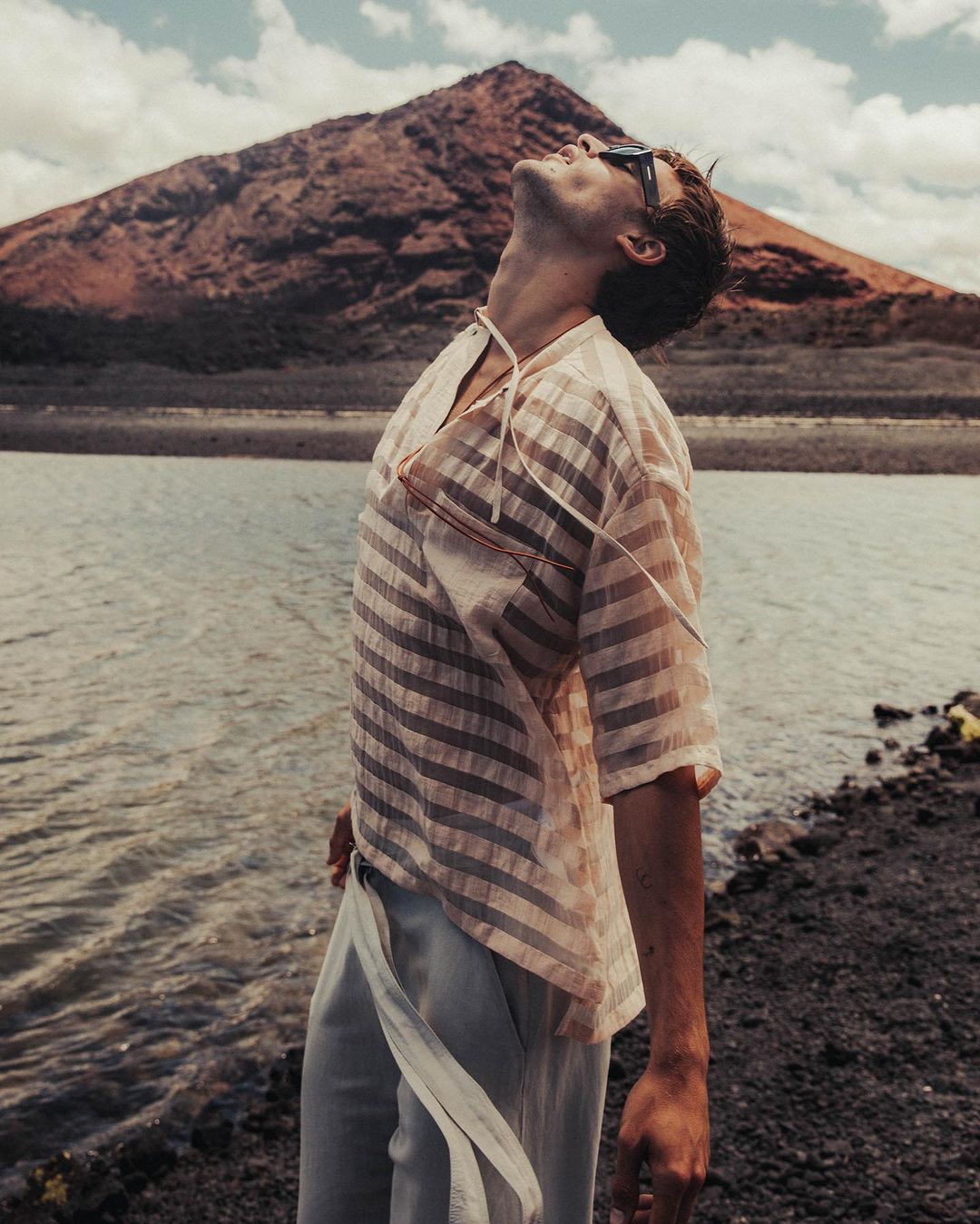 A man in a sheer, striped shirt and light pants stands near a lake with a reddish mountain in the background under a cloudy sky.