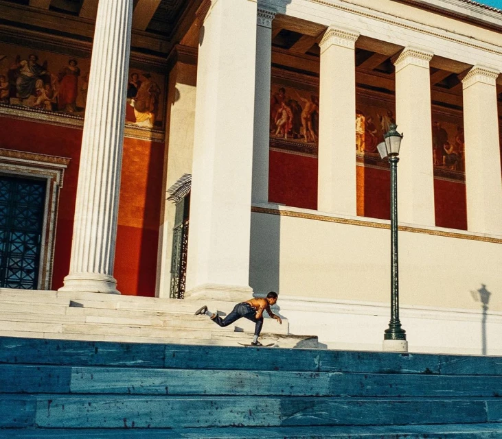 An adult rides a skateboard down a concrete staircase in front of an ornate stone building with columns on a sunny day.