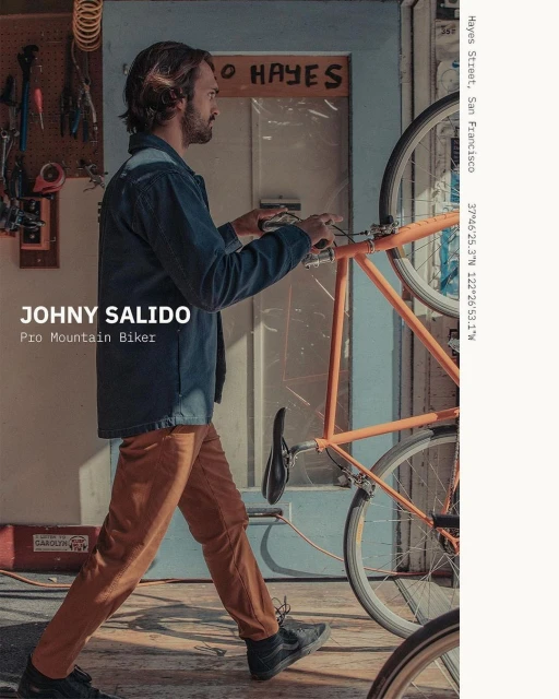Man in blue jacket walks with orange bicycle in a workshop setting, bathed in natural light, displaying an active lifestyle and urban scene.