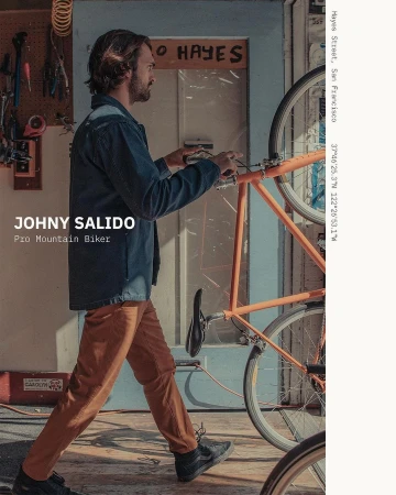 Man in blue jacket walks with orange bicycle in a workshop setting, bathed in natural light, displaying an active lifestyle and urban scene.