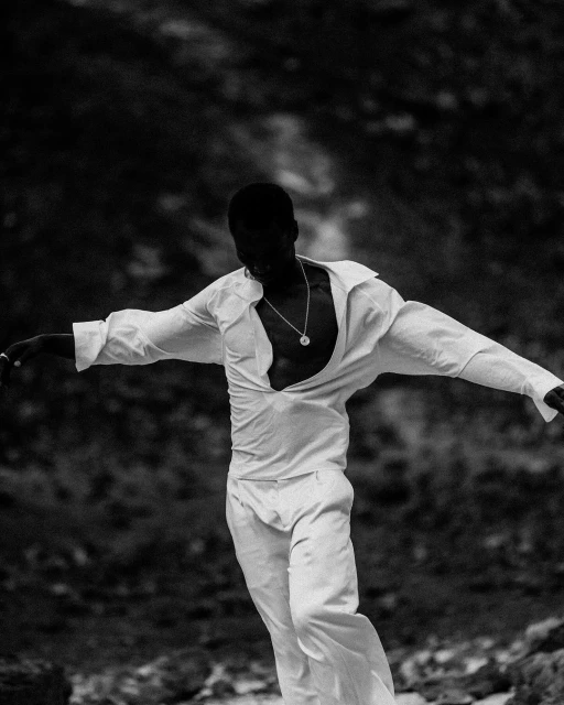 Monochrome image of a Black male model in an unbuttoned white shirt and pants striking a pose against a dark, rocky background.
