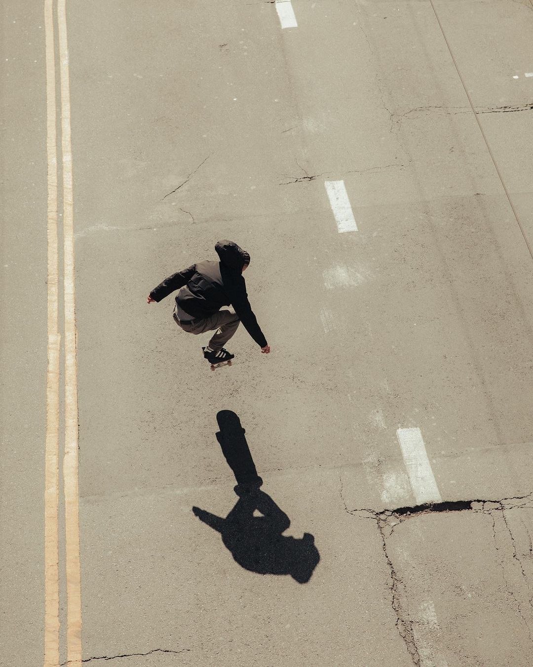 Young skateboarder in mid-air on an empty street, captured from an overhead angle, with the subject's shadow visible on the grey concrete.